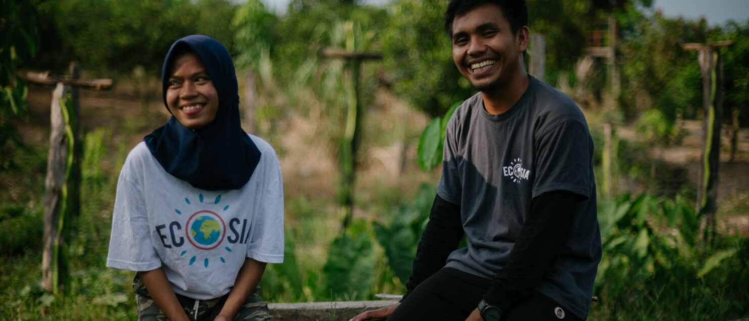 Eine junge Frau in einem Ecosia-T-Shirt und ein junger Mann sitzen nebeneinander strahlend auf einem Betonbecken. Hinter ihnen sieht man die Umrisse junger Bäume.